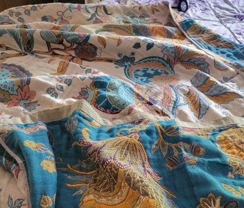 A close-up of a patterned quilt or blanket with a teal blue border and a multicolor paisley and floral design on an off-white background.